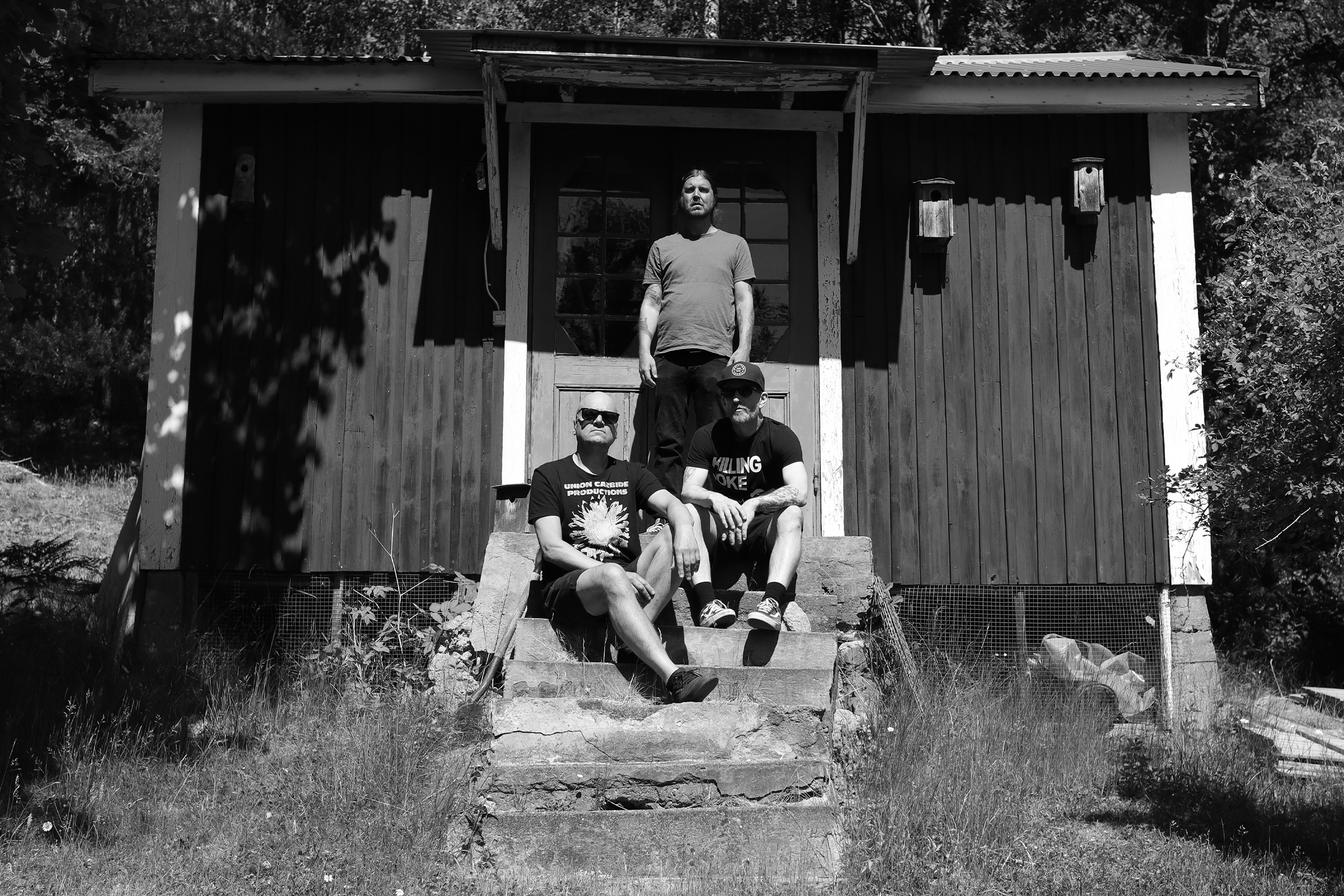 Black and white photo of Haystack outdoors at Studio Underjord: Ulf sitting on steps, Jonas seated higher up, and Patrik standing by the wooden double door, surrounded by nature and traditional Swedish architecture, photographed by Joona Hassinen.