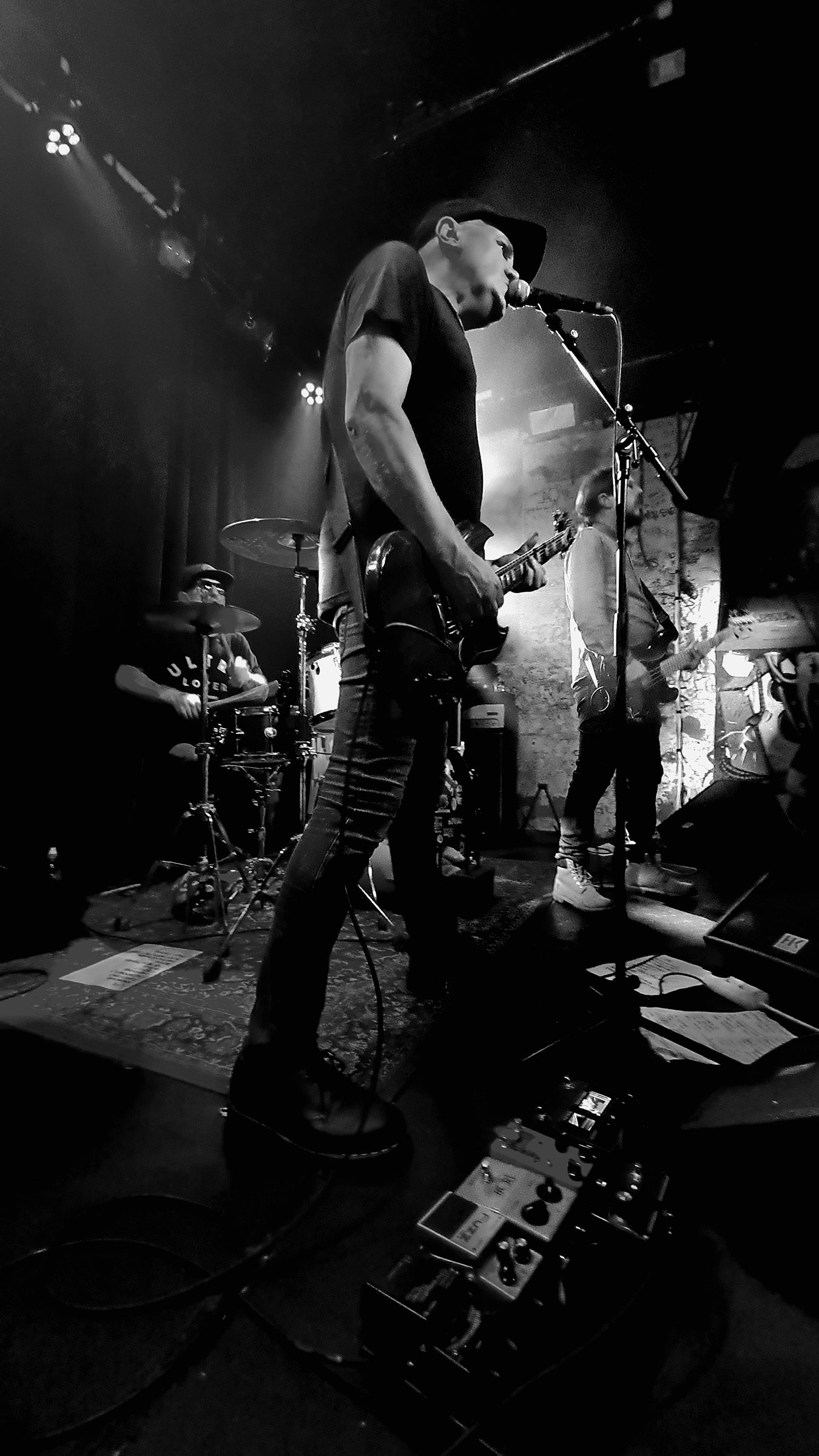 Black and white vertical photo of Haystack's Ulf 'Uffe' Cederlund with his guitar, pedals prominently at a 45-degree angle in the foreground, Jonas Lundberg on drums, and Patrik bisected by the mic stand, live at Kafé 44 in Stockholm, Sweden, on October 14, 2023.