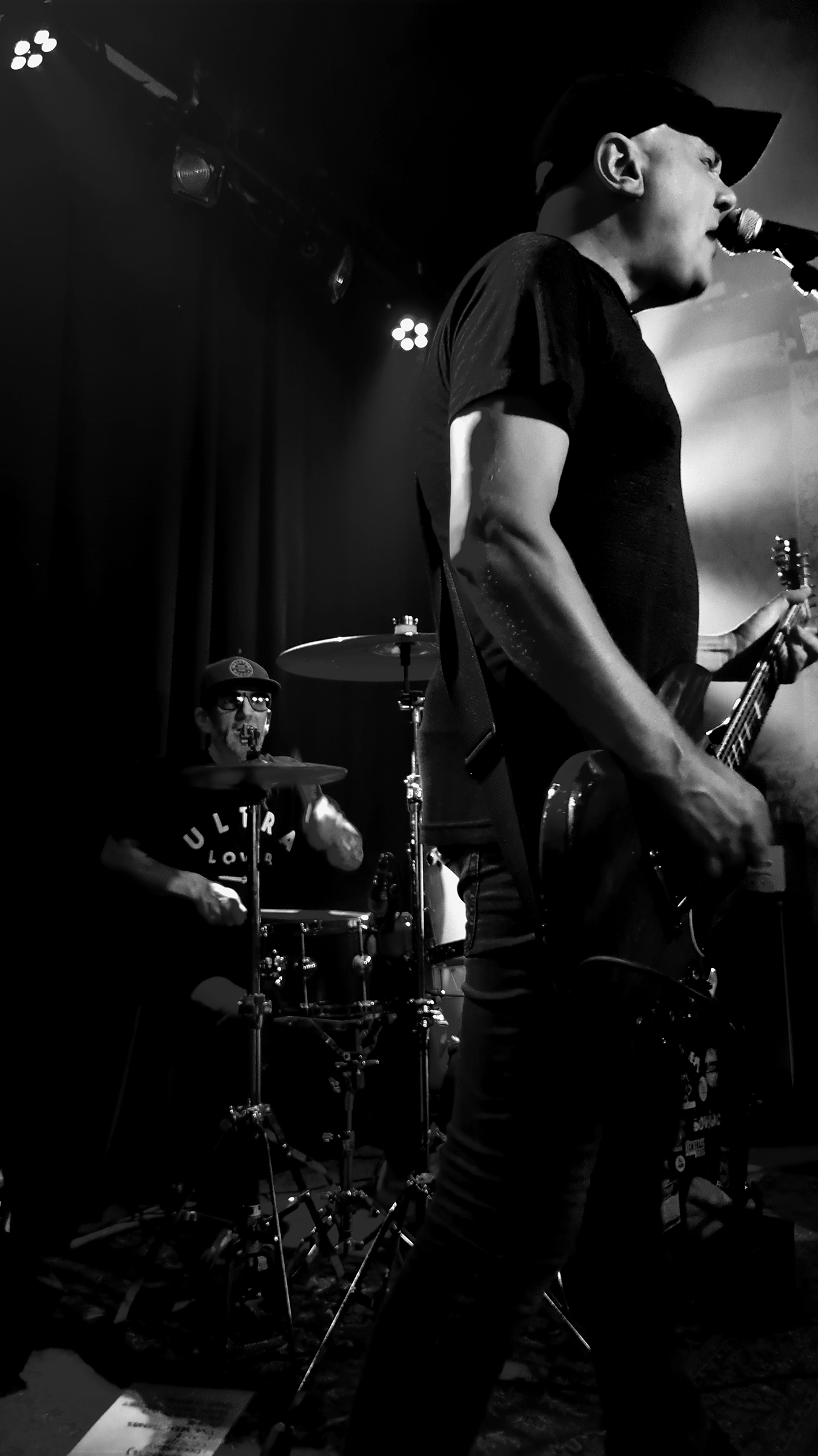 Black and white vertical photo showing Haystack's Ulf Cederlund nearly full-figure playing guitar and singing, with drummer Jonas Lundberg in the background, live at Kafé 44 in Stockholm, on October 14, 2023.