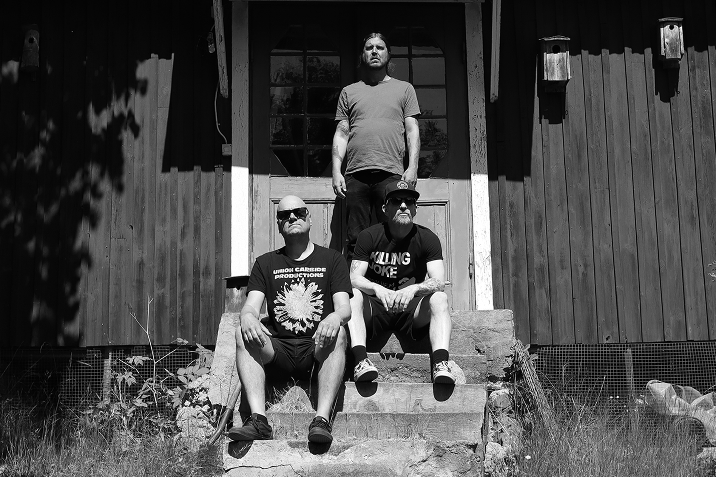 Black and white close-up photo of Haystack members at Studio Underjord: Ulf facing the camera in Union Carbide Productions shirt and sunglasses, Jonas in a Killing Joke t-shirt and baseball cap, Patrik partially visible behind them, all under midday sun shadows.