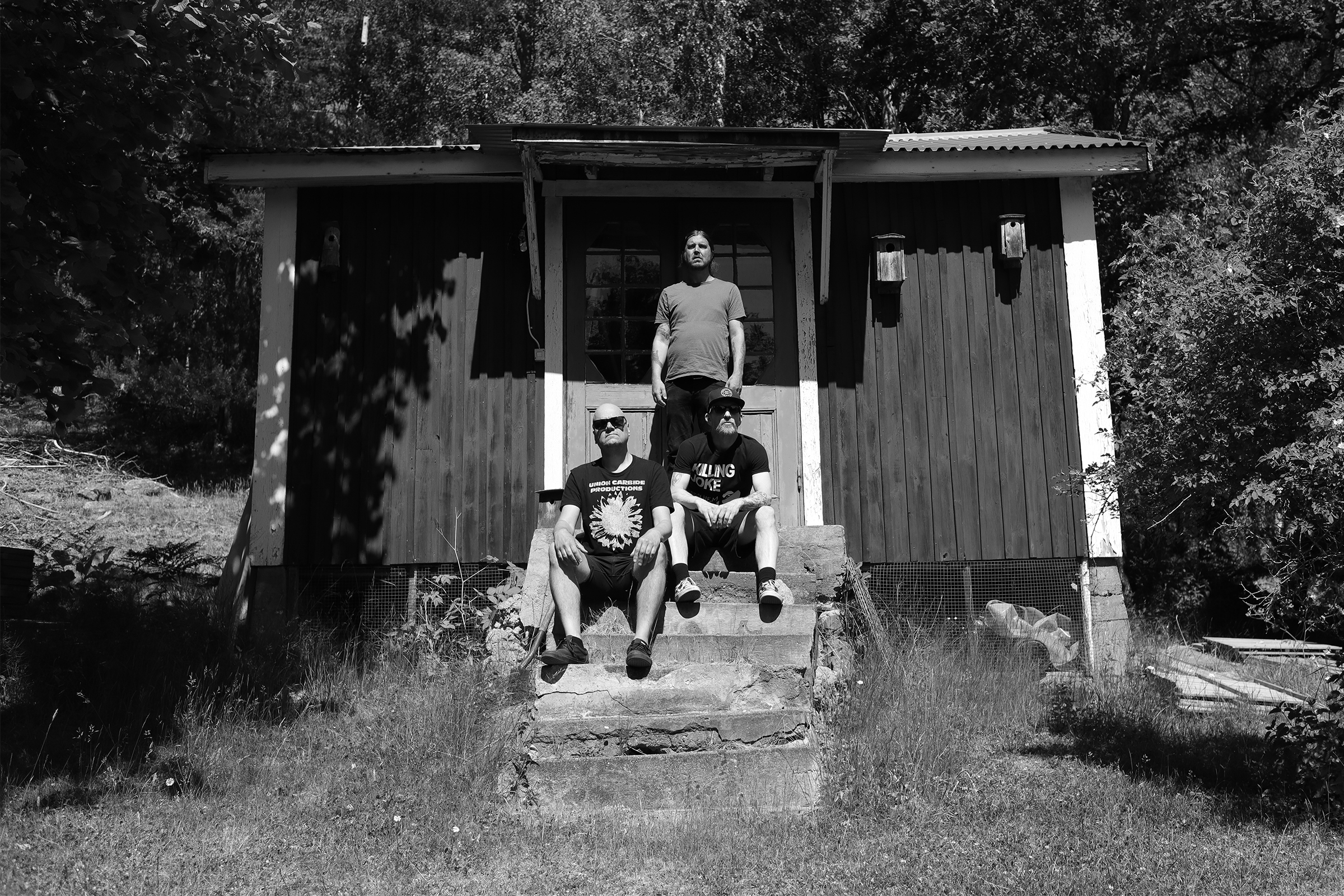 Black and white horizontal photo of Haystack at Studio Underjord: Ulf, Jonas, and Patrik in a relaxed pose with a lush backdrop of trees and bushes, captured by Joona Hassinen.