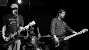 Black and white photo of Haystack performing live at Gamla Enskede Bryggeri, with Ulf Cederlund on guitar and vocals, Jonas Lundberg on drums, and Patrik Thorngren on bass.
